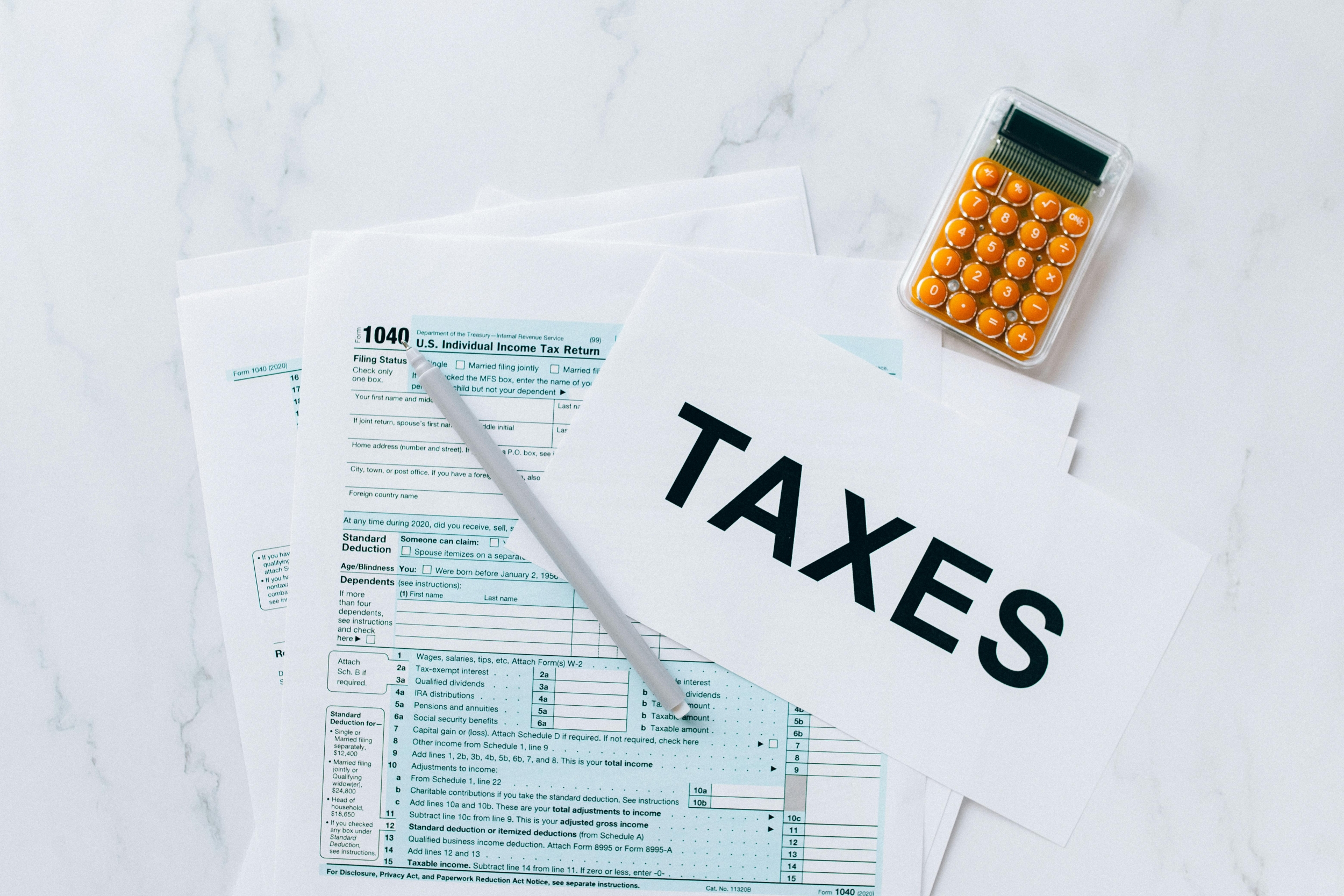 Flat-lay of a U.S. Form 1040 tax return with a pen, an orange calculator, and a card labeled "TAXES" on a marble surface.